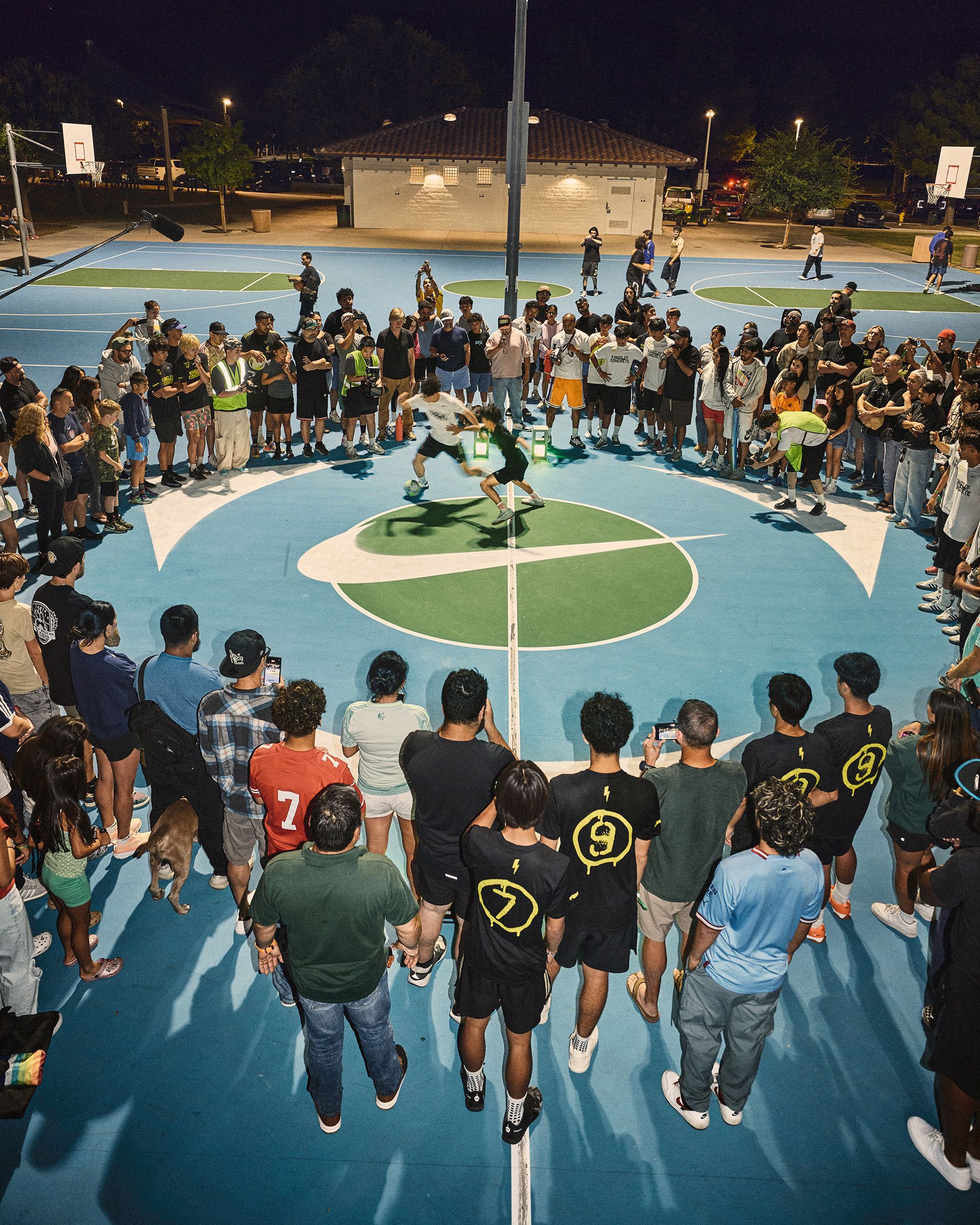A large crowd surrounds two people playing basketball on an outdoor court at night, with spectators closely watching and some filming with their phones.