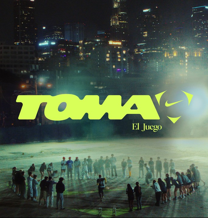 A large group of people forms a circle in an outdoor urban area at night, with city buildings lit up in the background. The text "TOMA El Juego" is prominently displayed.