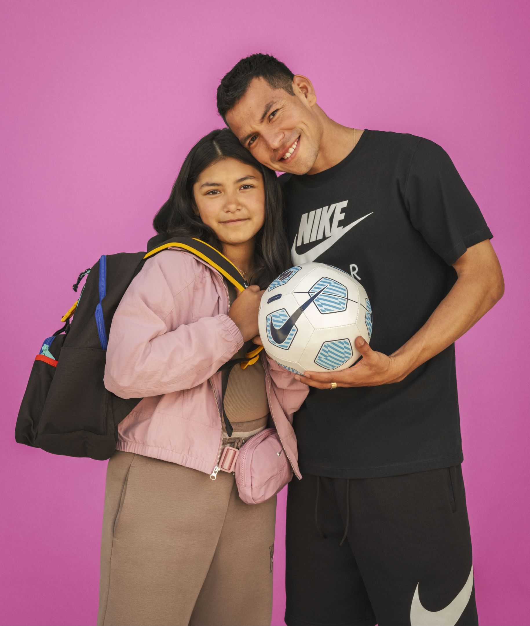 A man and a girl stand together against a pink background; the girl holds a backpack, and the man holds a soccer ball. Both are smiling.