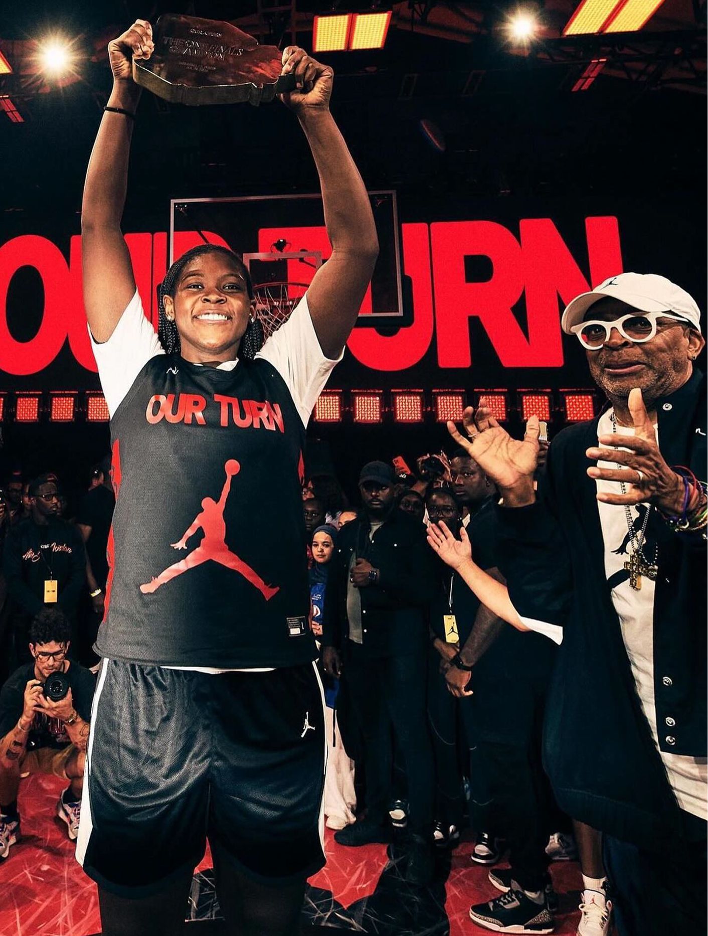 A basketball player holds up a trophy in celebration, wearing a shirt with the Jordan logo, while a man applauds nearby. Red "OUR TURN" text is displayed in the background.