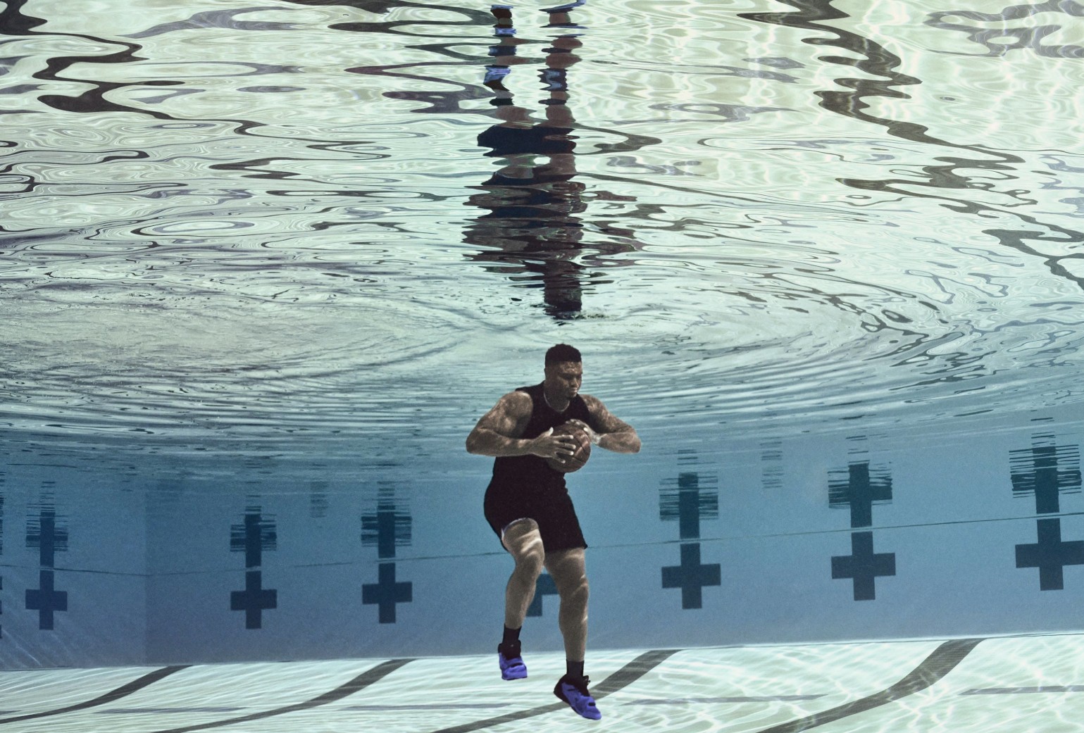 A person in athletic wear walks underwater while holding a football in a swimming pool, with their reflection visible on the water’s surface.