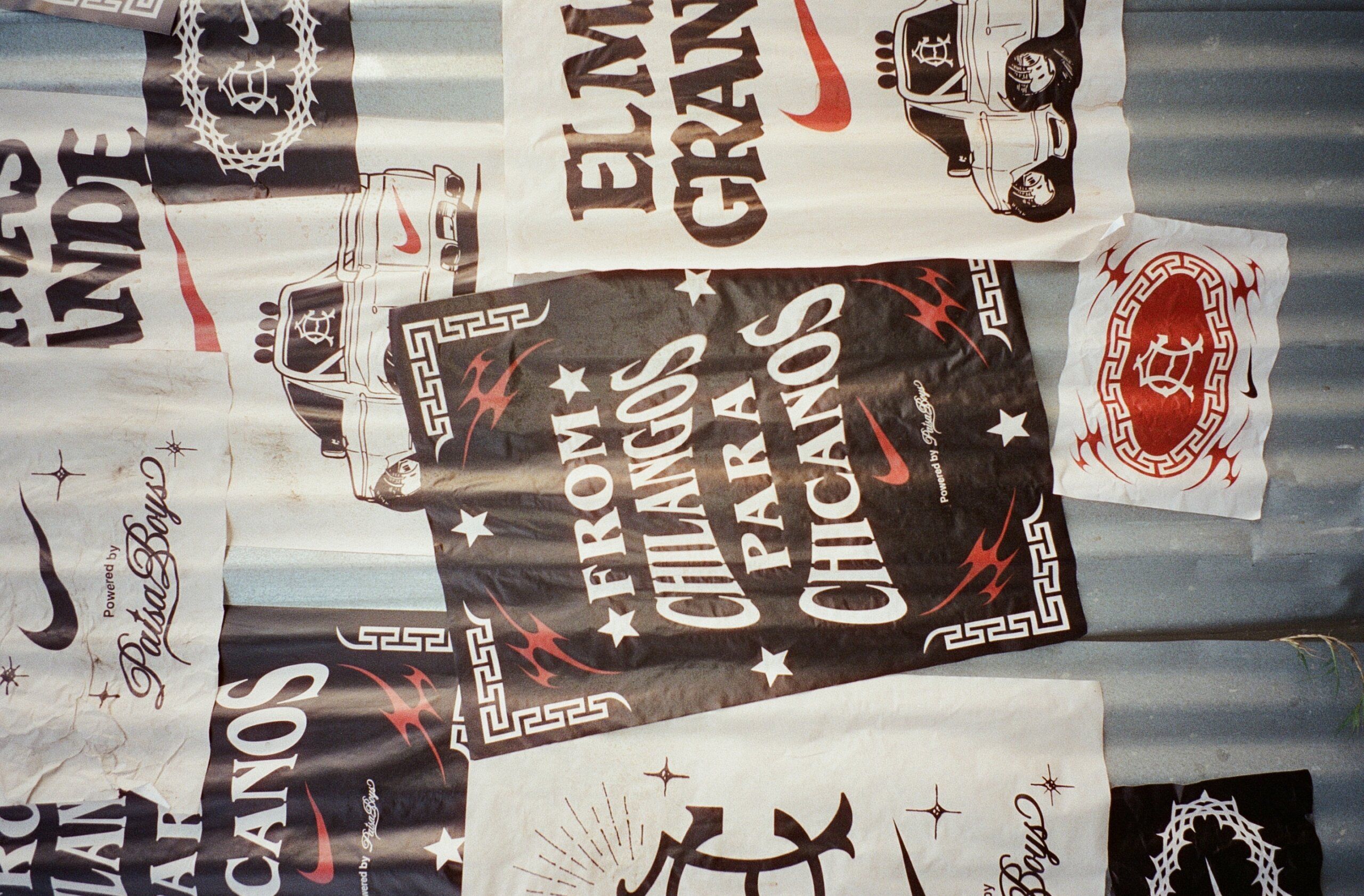 Assorted posters with bold graphics and text, including one that reads "FROM CHILANGOS PARA CHICANOS," are displayed on a corrugated metal wall.