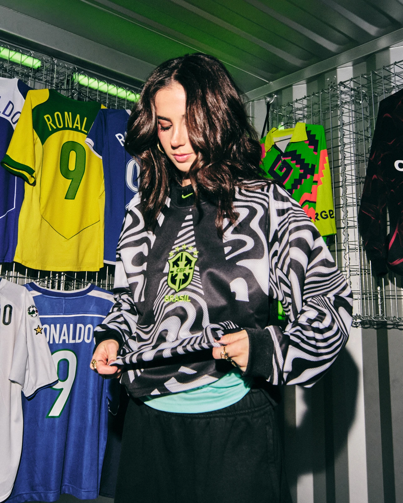 A woman stands in front of a wall displaying football jerseys, wearing a black and white patterned hoodie and looking down while adjusting her clothing.
