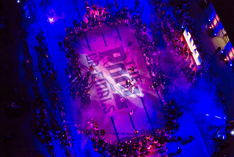 A crowd surrounds a stage with “THE ROAD TO FINALS” displayed in large letters, illuminated by purple and blue lights, viewed from above.