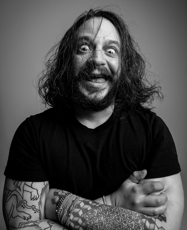 A man with long hair and tattoos on both arms stands with his arms crossed, wearing a black t-shirt and smiling widely in a black-and-white portrait.