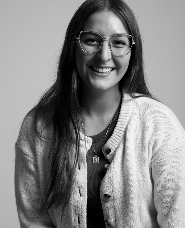 A young woman with long hair, wearing glasses, a cardigan, and layered necklaces, smiles at the camera in a black and white portrait.