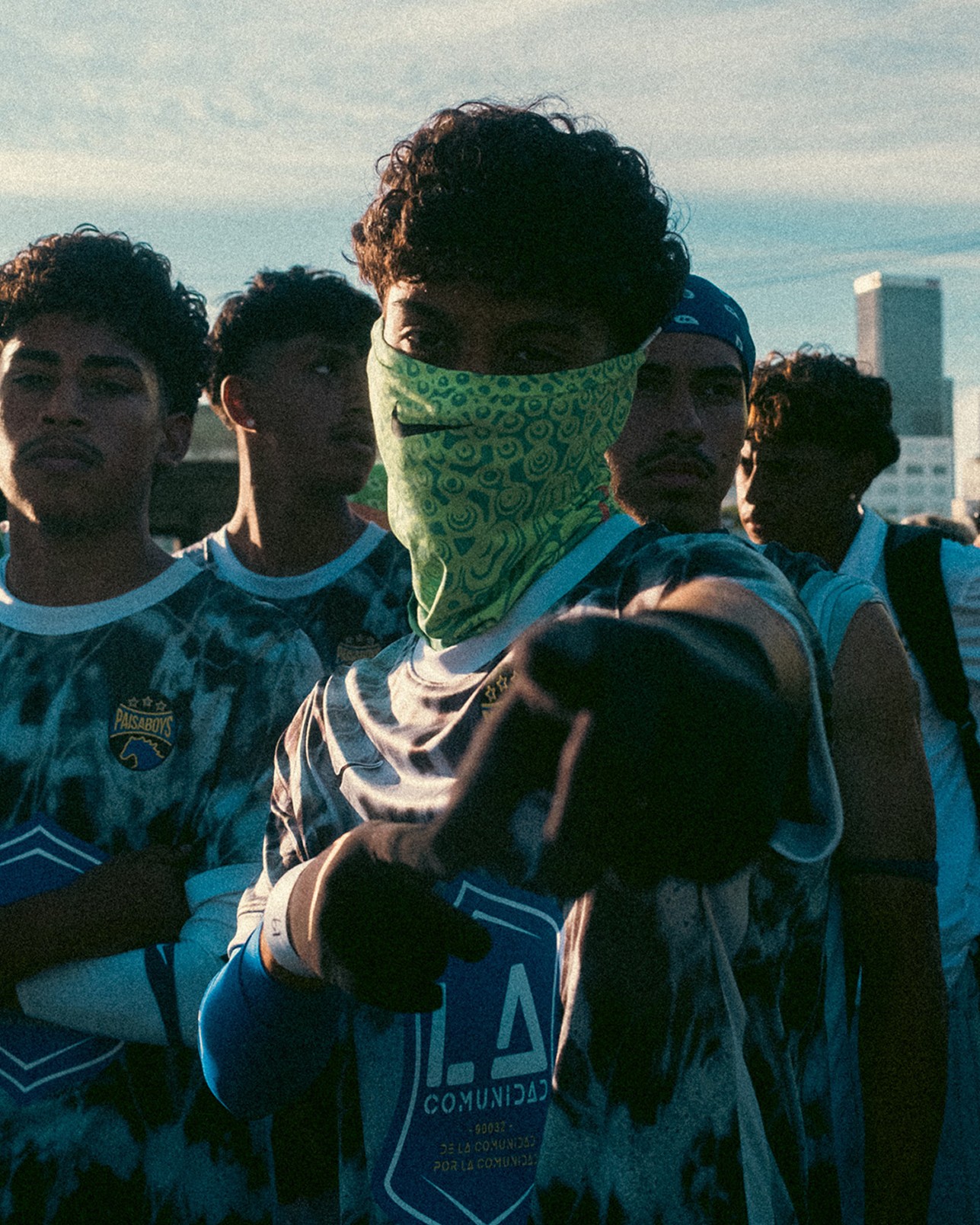 A group of young men in sports jerseys stand together outdoors; one in front wears a green bandana over his face and points towards the camera.