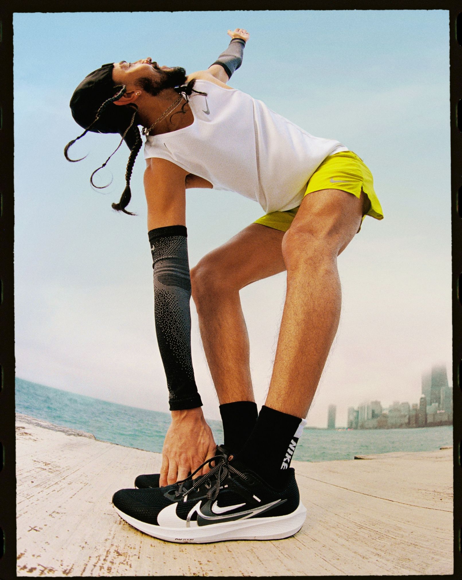 Man doing standing side angle stretch getting ready to run. Has two braids, beard, arm warmers, white tank, green shorts and black nike pegasus running shoes. He is on a dock in front of a body of water with cityscape on other side