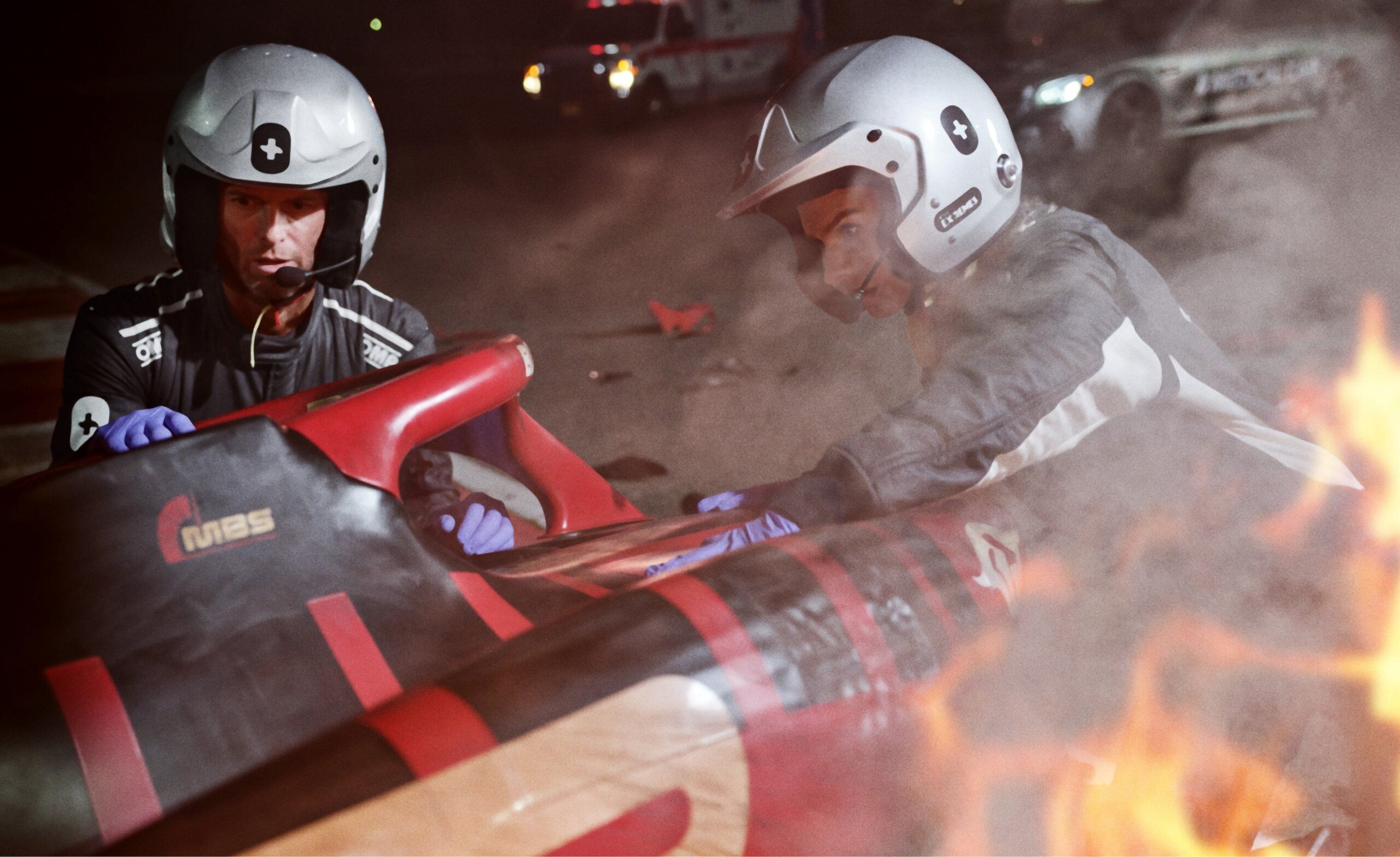 Two medical technicians with helmets and safety gear attend to F1 car driver in firey crash