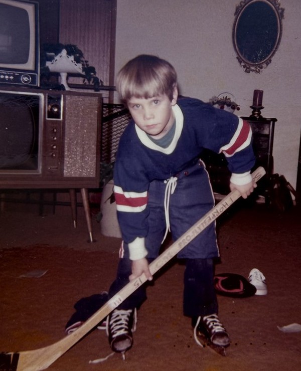 A young boy in hockey gear poses with a hockey stick indoors, standing on carpet in a living room with vintage furniture and a TV in the background.