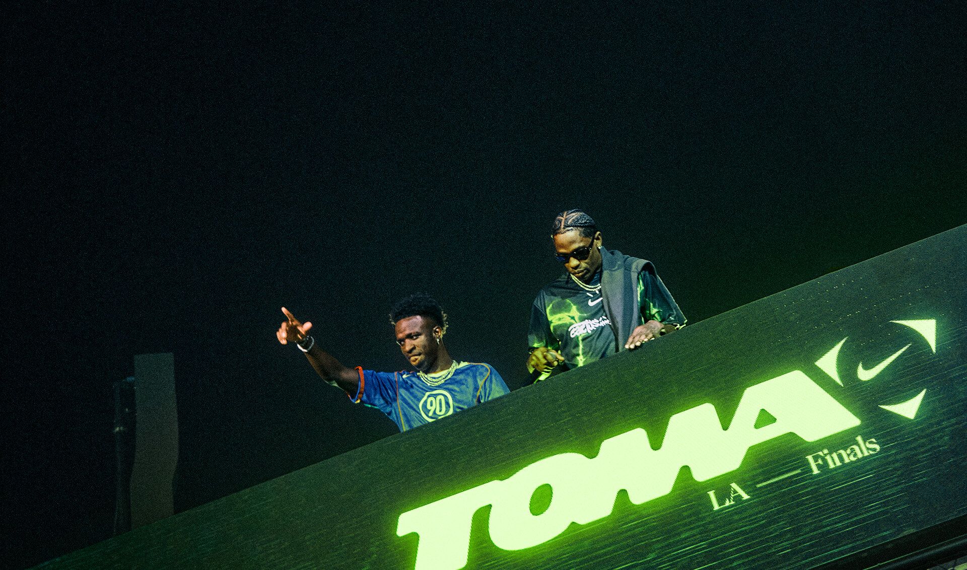 Two men stand behind a large illuminated sign that reads "TOMA El Juego LA Finals" at an elevated venue, with one pointing into the crowd.