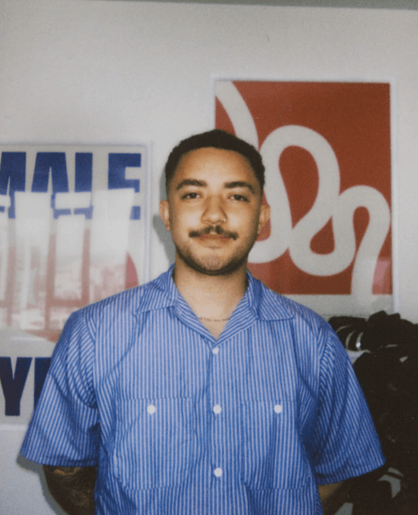 A person with short hair and a mustache stands indoors, wearing a blue striped shirt. Colorful posters are visible on the wall behind them.