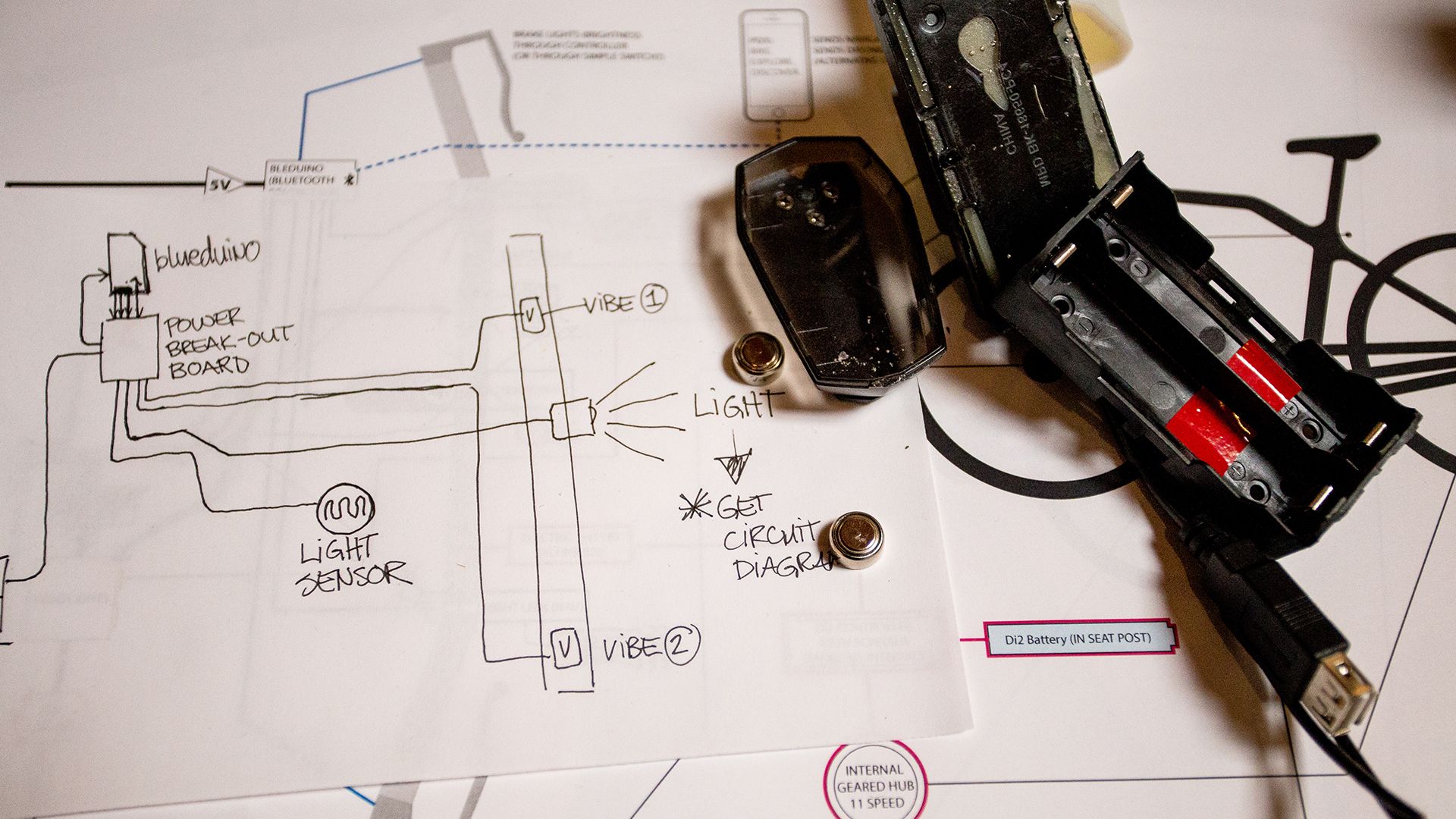 A cluttered desk with a hand-drawn electronic circuit diagram, a disassembled device, batteries, and various components.