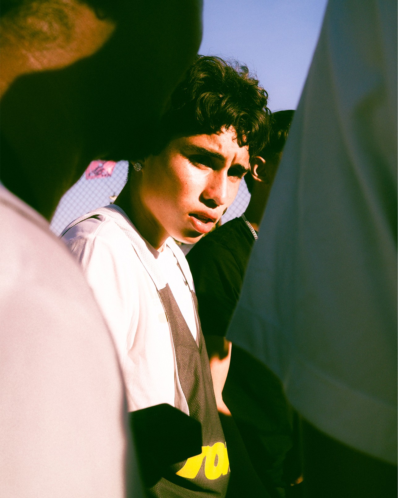 A young person in a white shirt stands outdoors, looking towards the camera, partially framed by other people in the foreground.
