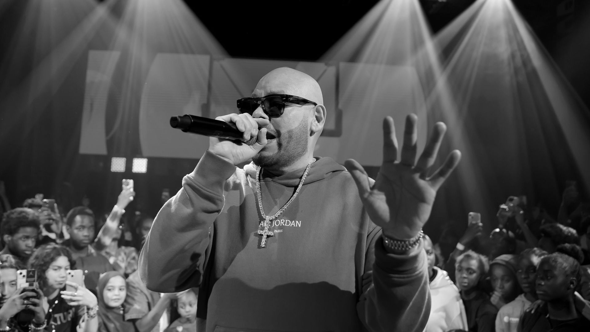 A man wearing sunglasses and a hoodie performs on stage with a microphone, raising one hand, surrounded by a crowd.