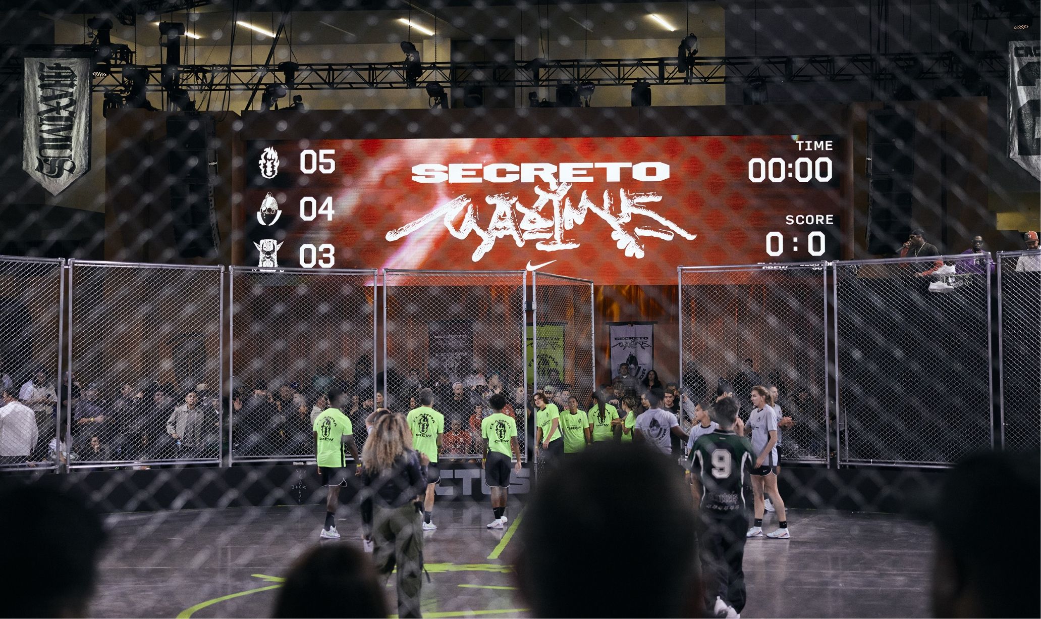 Indoor soccer match with players wearing neon green and black jerseys. A large scoreboard displays the word "SECRETO" against a crowd-filled background.