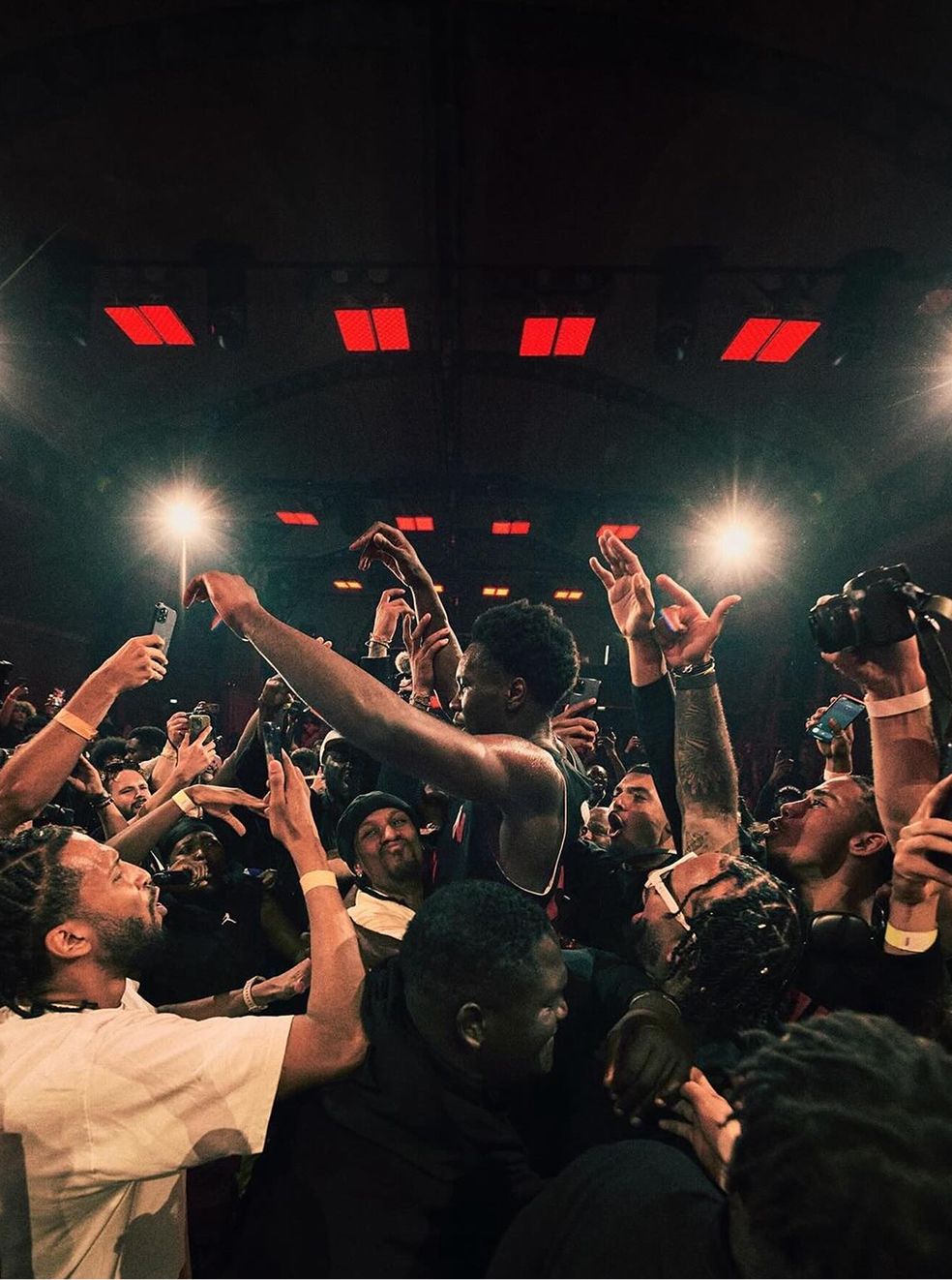 A basketball player is surrounded by an excited crowd of fans and photographers, many holding up phones and cameras to capture the moment.