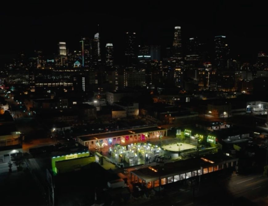 An illuminated outdoor boxing ring and surrounding buildings at night with a city skyline in the background.