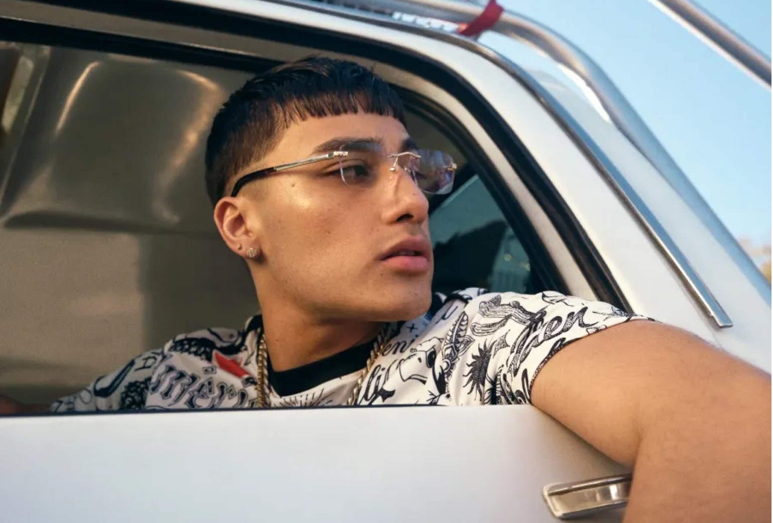 A person wearing clear glasses and a patterned shirt looks to the side while sitting in a car with the window down.