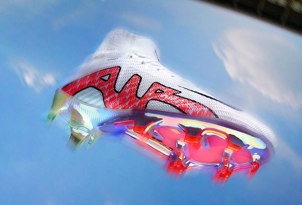 A football boot with a white upper, red and black design accents, and a translucent sole featuring red, orange, and purple studs, photographed against a blue sky background.