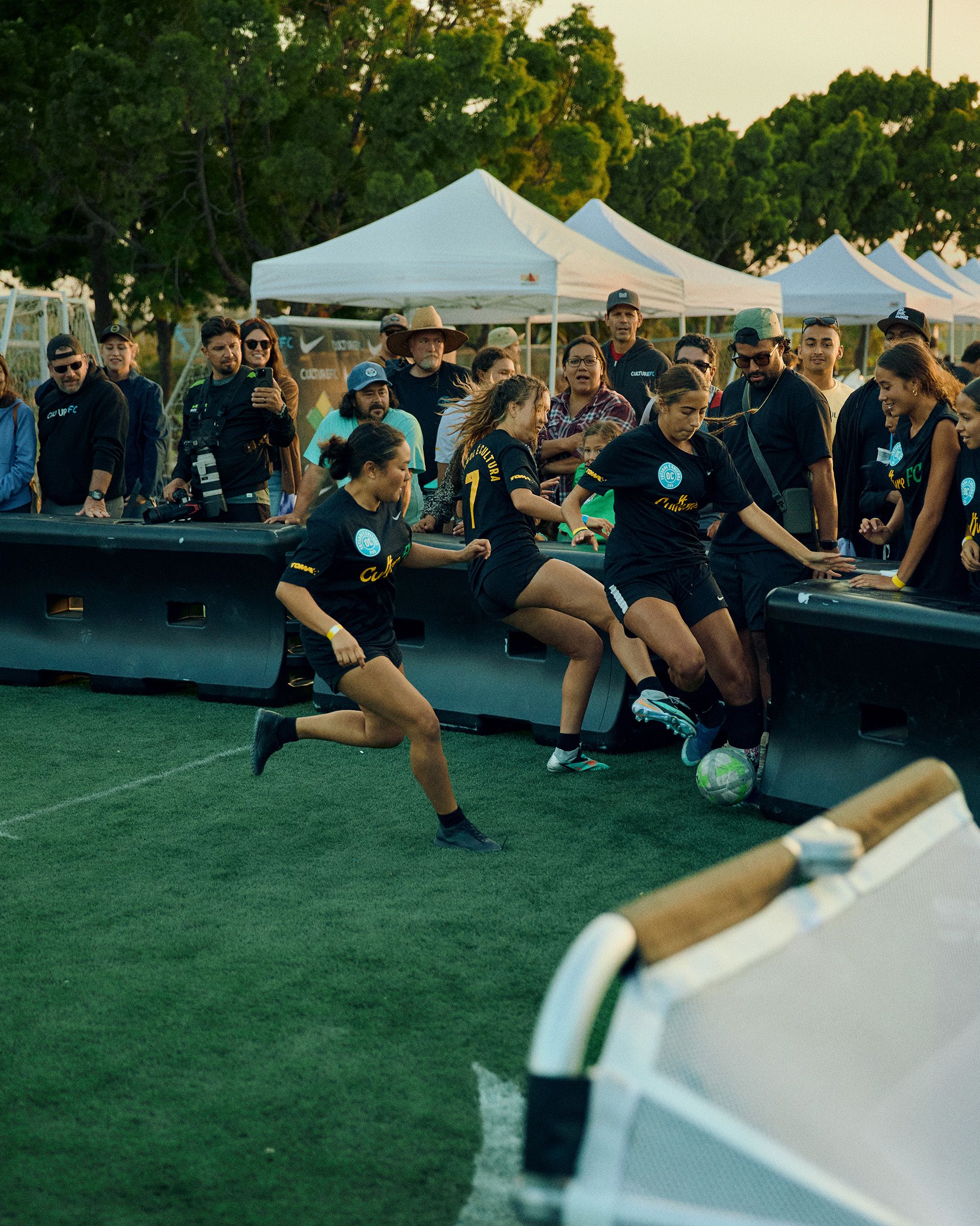 Three female soccer players in black uniforms compete for the ball near the sideline, while spectators and photographers watch from behind a barrier at an outdoor event.
