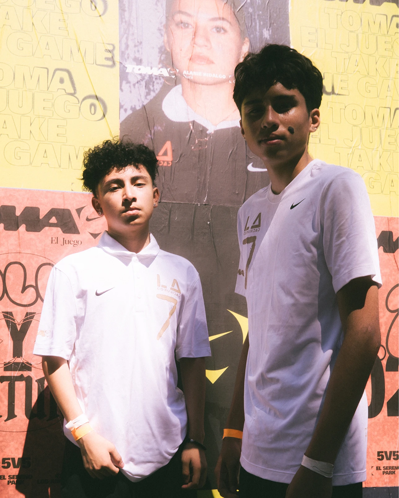Two teenage boys wearing white Nike shirts stand in front of a colorful poster-covered wall, looking at the camera.