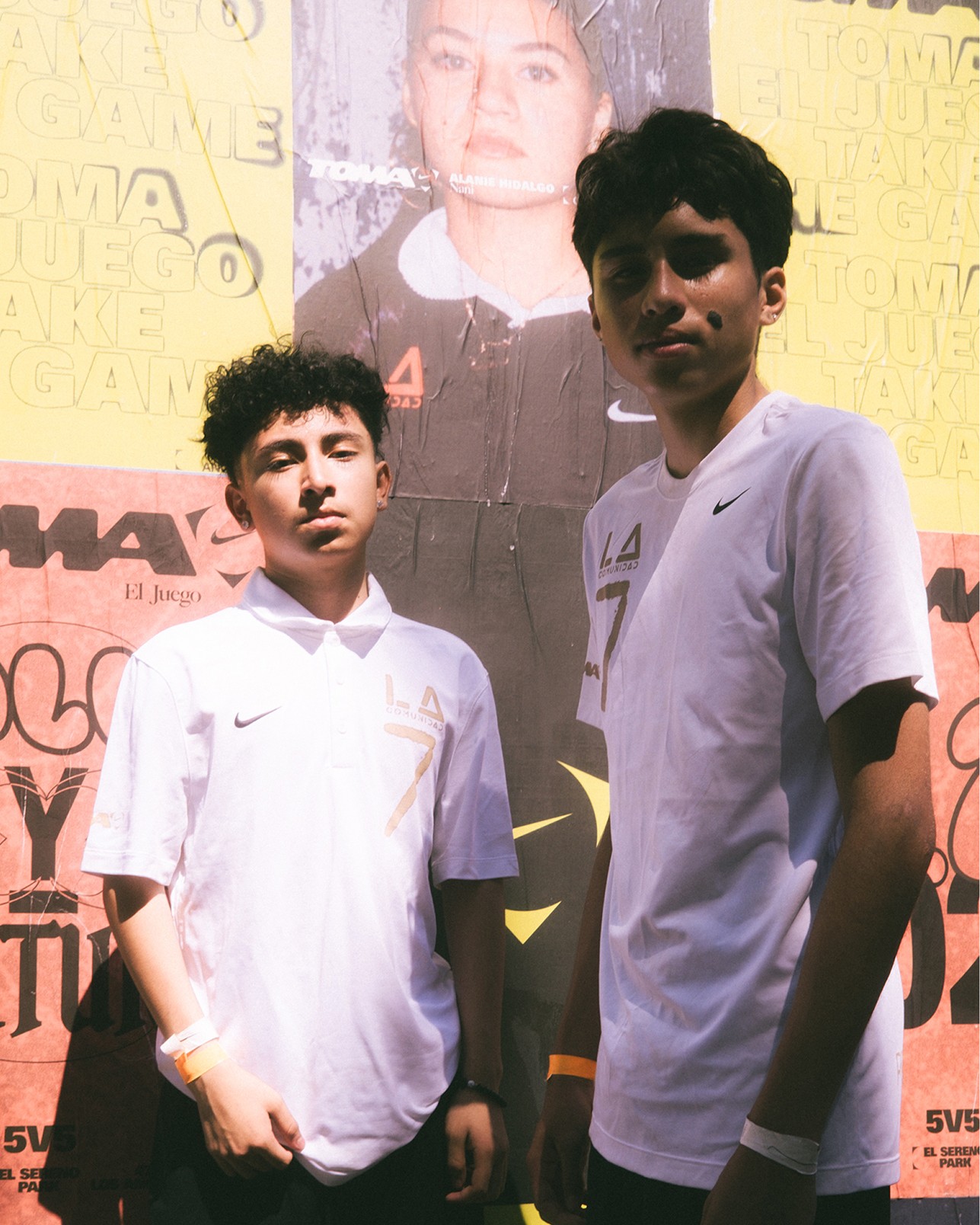 Two teenage boys wearing white Nike shirts stand in front of a colorful poster-covered wall, looking at the camera.
