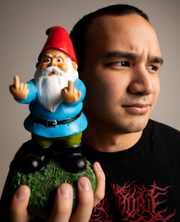 A person holds a garden gnome figurine with both hands raised, showing its middle fingers. The person looks to the side with a neutral expression.