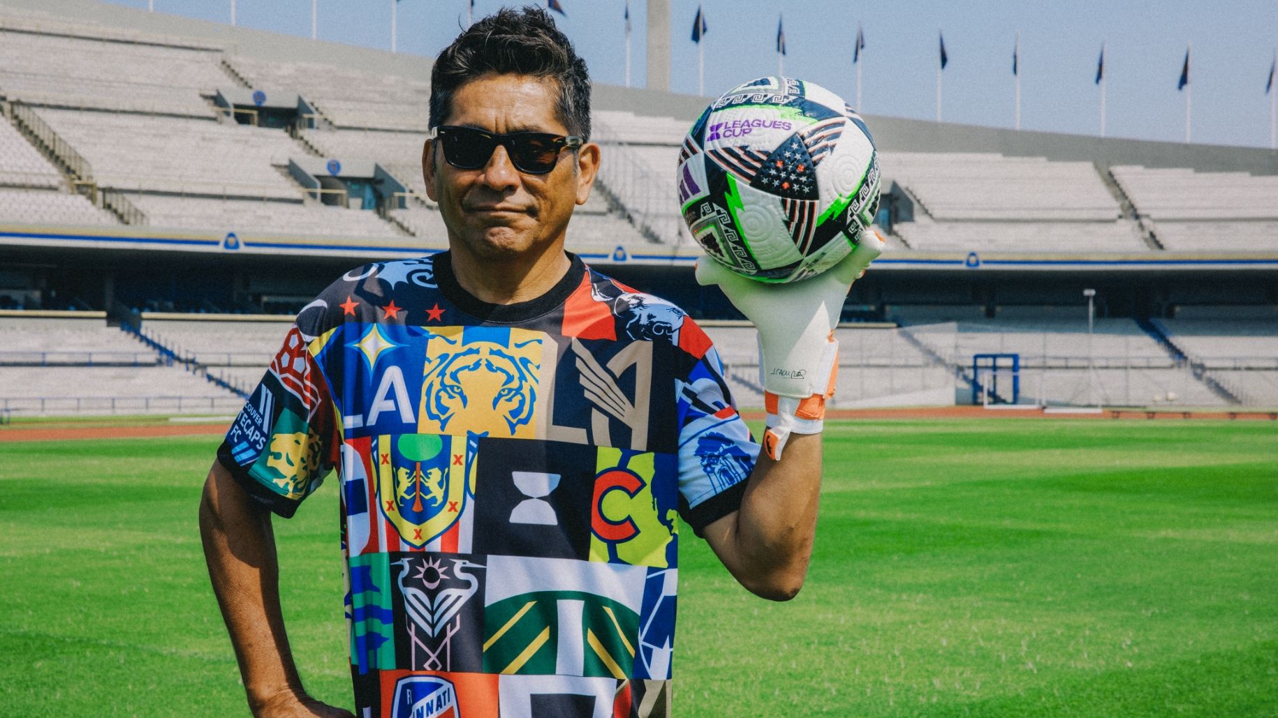 A person wearing sunglasses and a colorful shirt stands on a soccer field, holding a soccer ball.