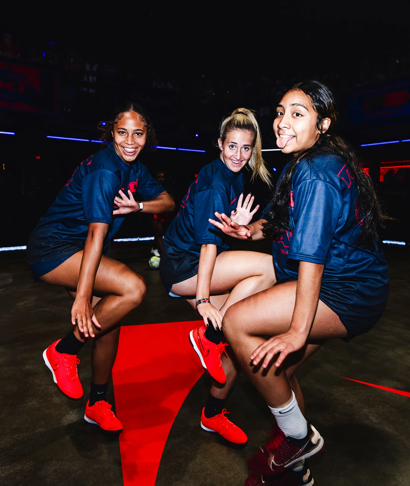Three women in matching navy shirts and red sneakers pose in a crouched position on an indoor court, smiling at the camera.