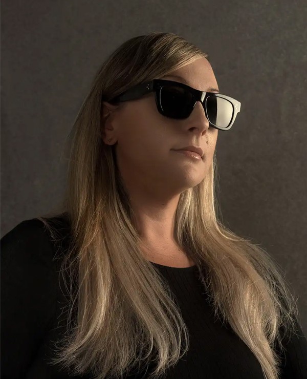 A person with long blonde hair wearing black sunglasses and a black top poses against a plain, neutral background.
