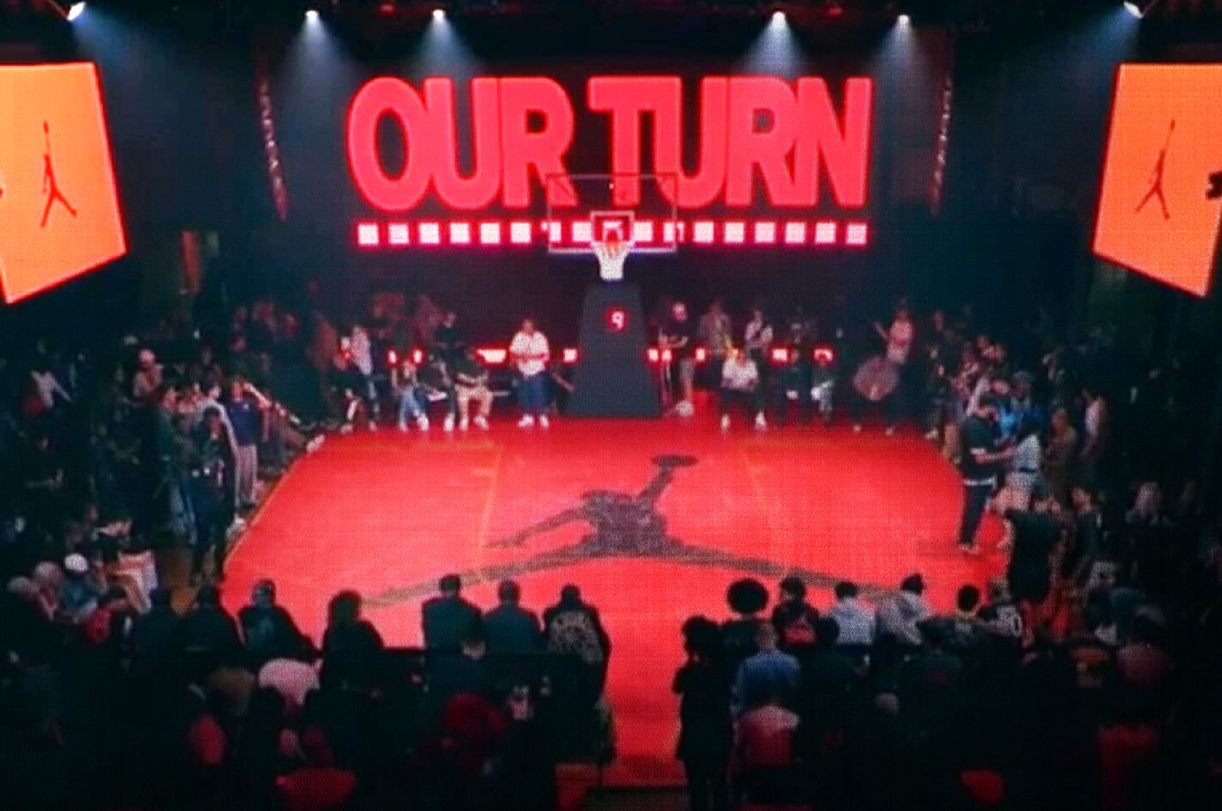 Indoor basketball court event with a red floor and large "OUR TURN" sign above the hoop; audience surrounds the court, and a Jordan logo is at center court.