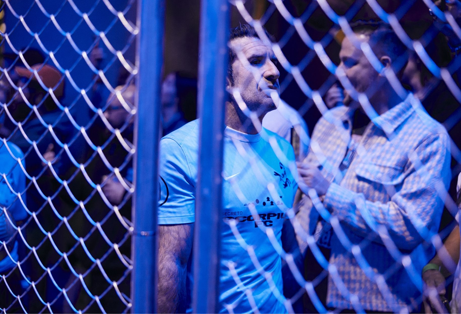 A man wearing a light blue shirt stands behind a chain-link fence, surrounded by people.