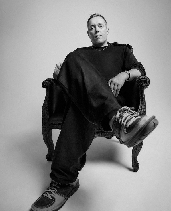 A person sits in a decorative armchair, dressed in dark clothing and sneakers, looking directly at the camera in a simple studio setting.