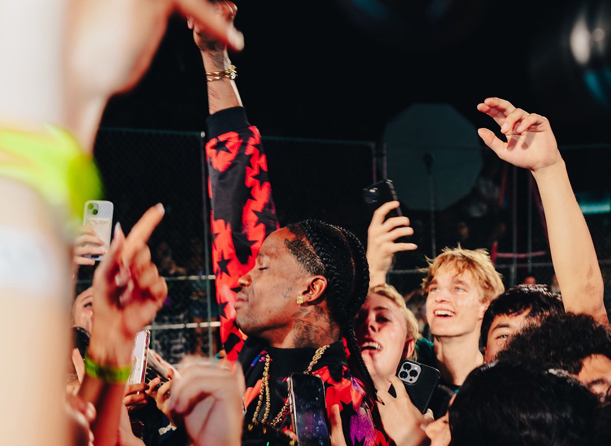 A performer in a red and black outfit raises an arm while surrounded by cheering fans with phones at an outdoor event.