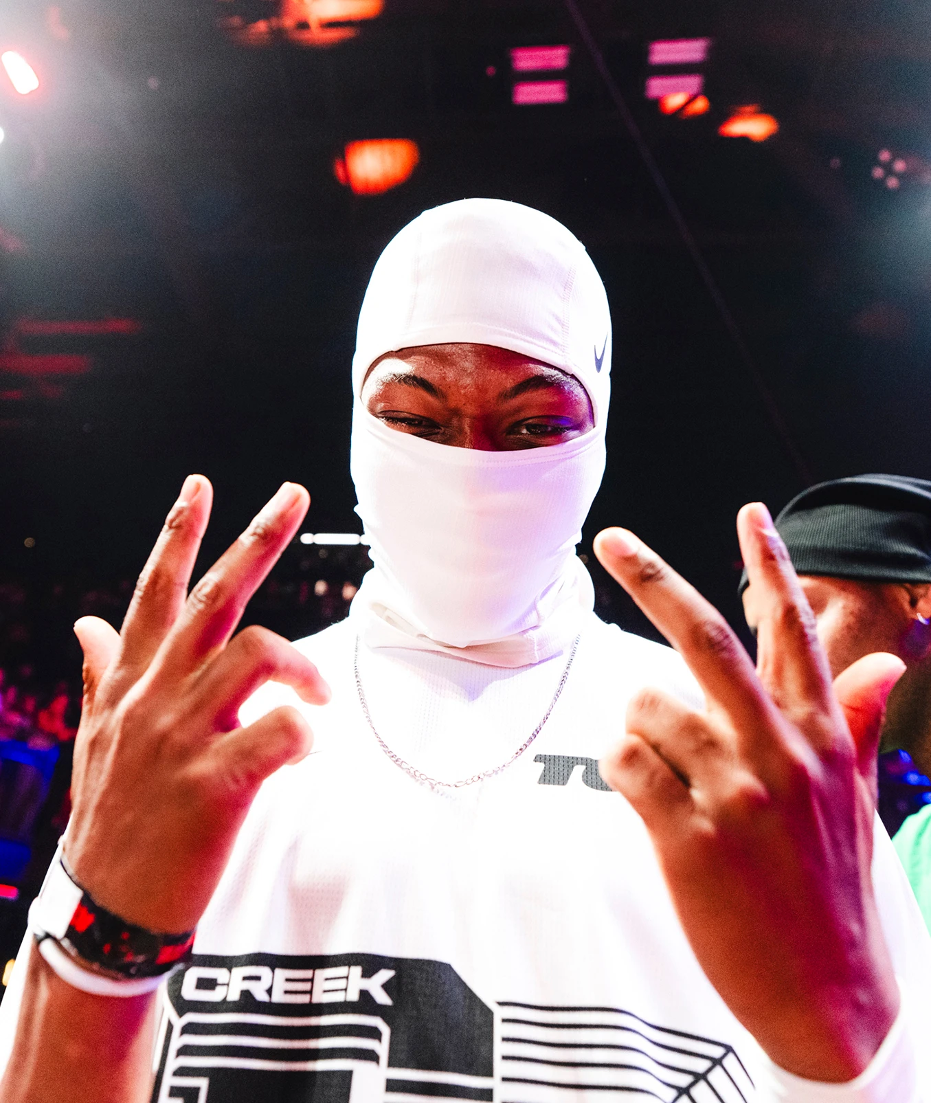 Person wearing a white balaclava and white shirt, posing with hands raised and fingers spread. The background is dimly lit with colorful lights.