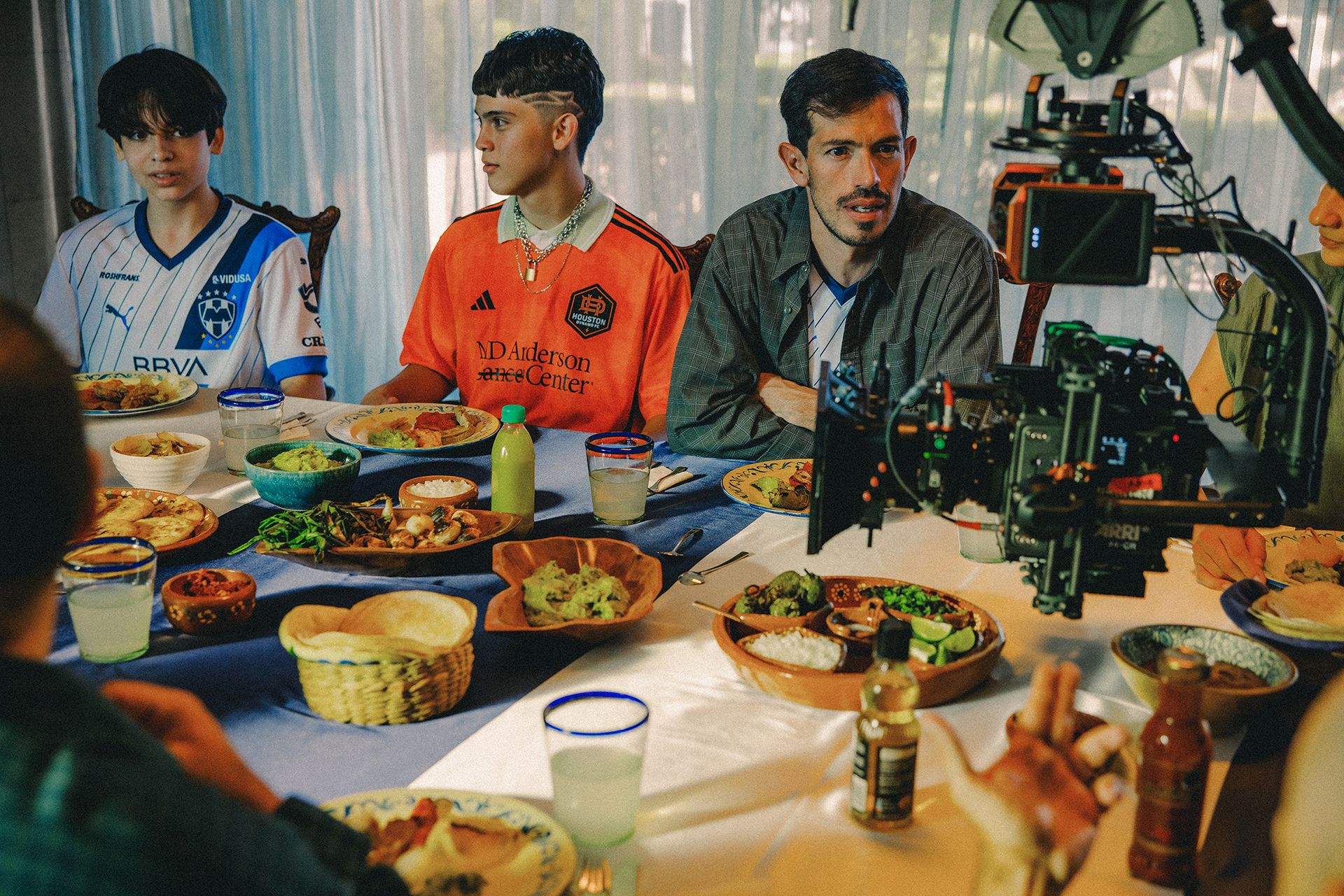 Three people are seated at a dining table with various dishes and drinks, while a video camera on a tripod records the scene.