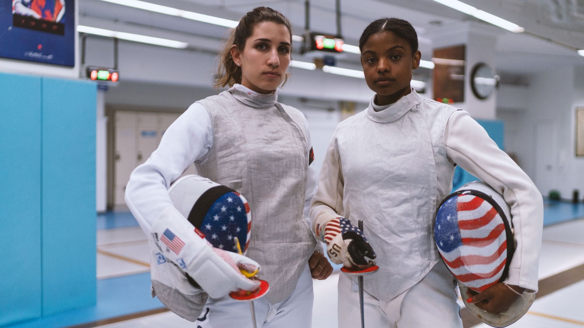 2 women fencers posing together in full protective gear with helmets off