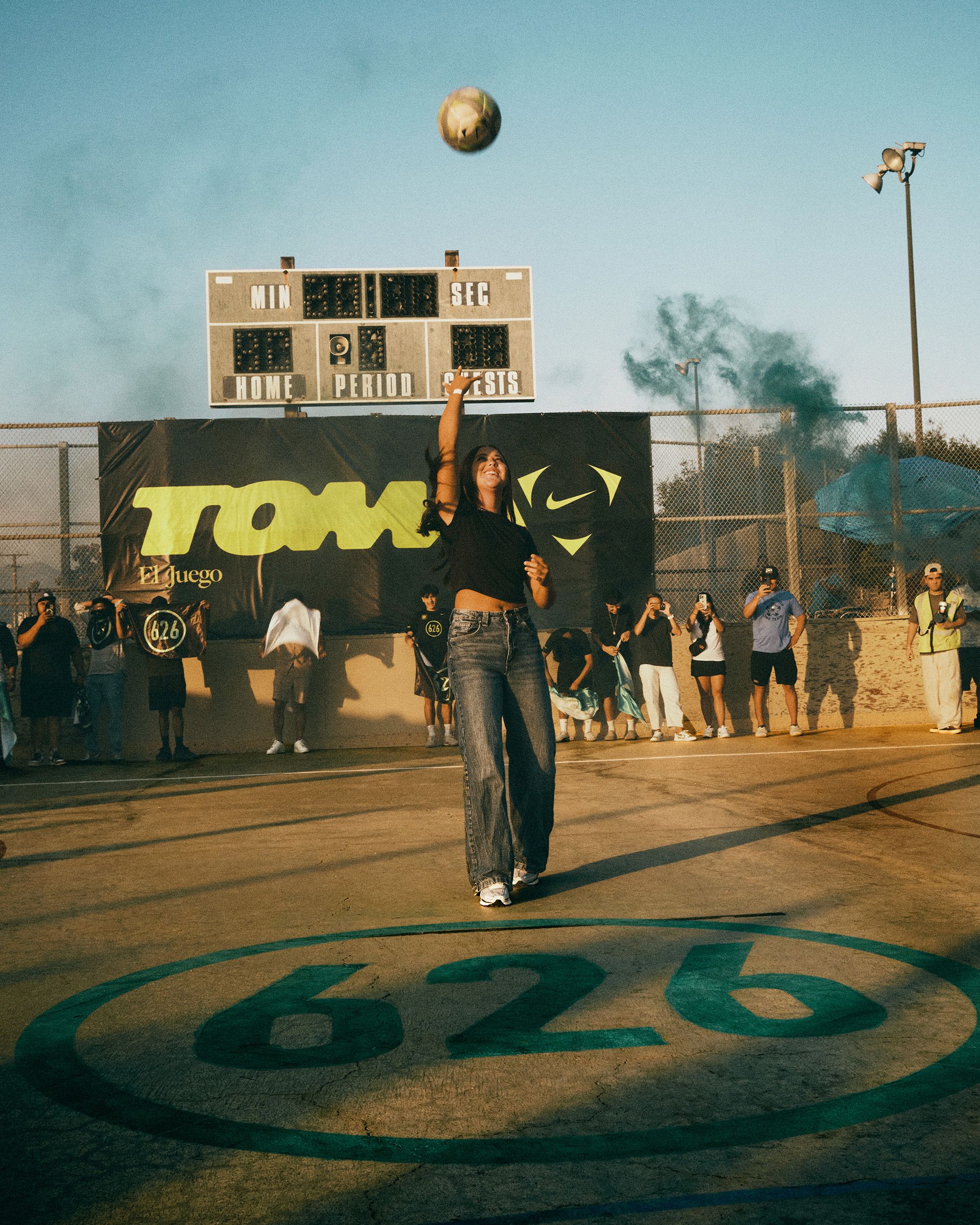 A woman stands on a basketball court with “626” painted on the ground, tossing a ball upward, while people and a scoreboard are visible in the background.