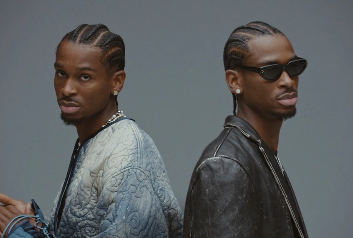 Two men with braided hair stand back-to-back; one wears a quilted jacket, the other wears sunglasses, earrings, and a black leather jacket, both facing slightly away from the camera.