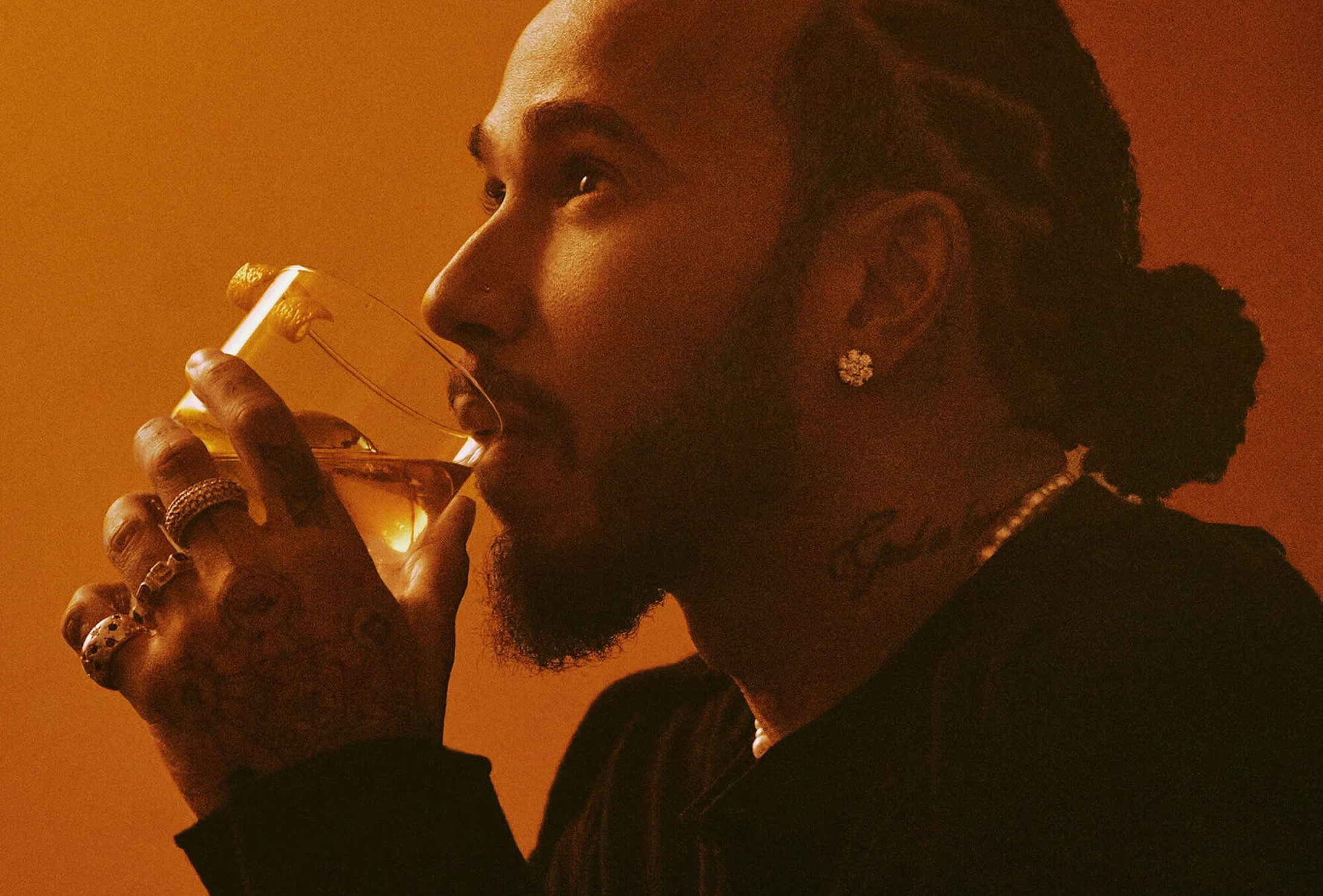 A man with braided hair and tattoos on his hand drinks from a glass, wearing earrings and a dark shirt, against a warm orange background.