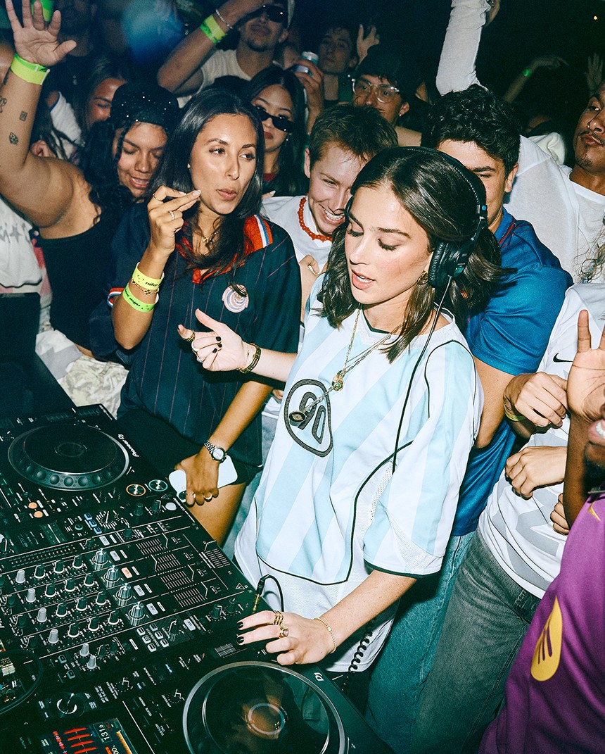A DJ mixes music at a crowded party, surrounded by people dancing and cheering. DJ equipment and turntables are visible in the foreground.