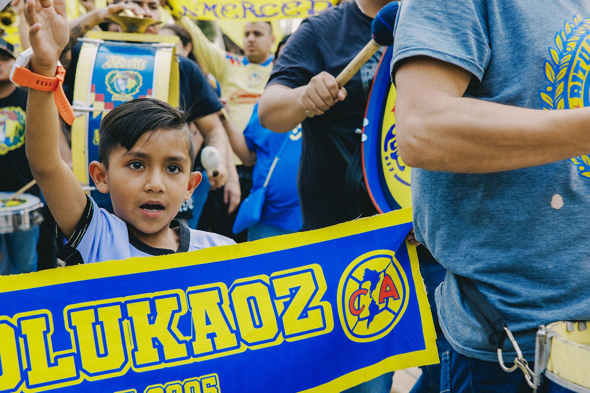 A young boy enthusiastically waves while surrounded by people holding a large, colorful banner with bold text and logos during an event.