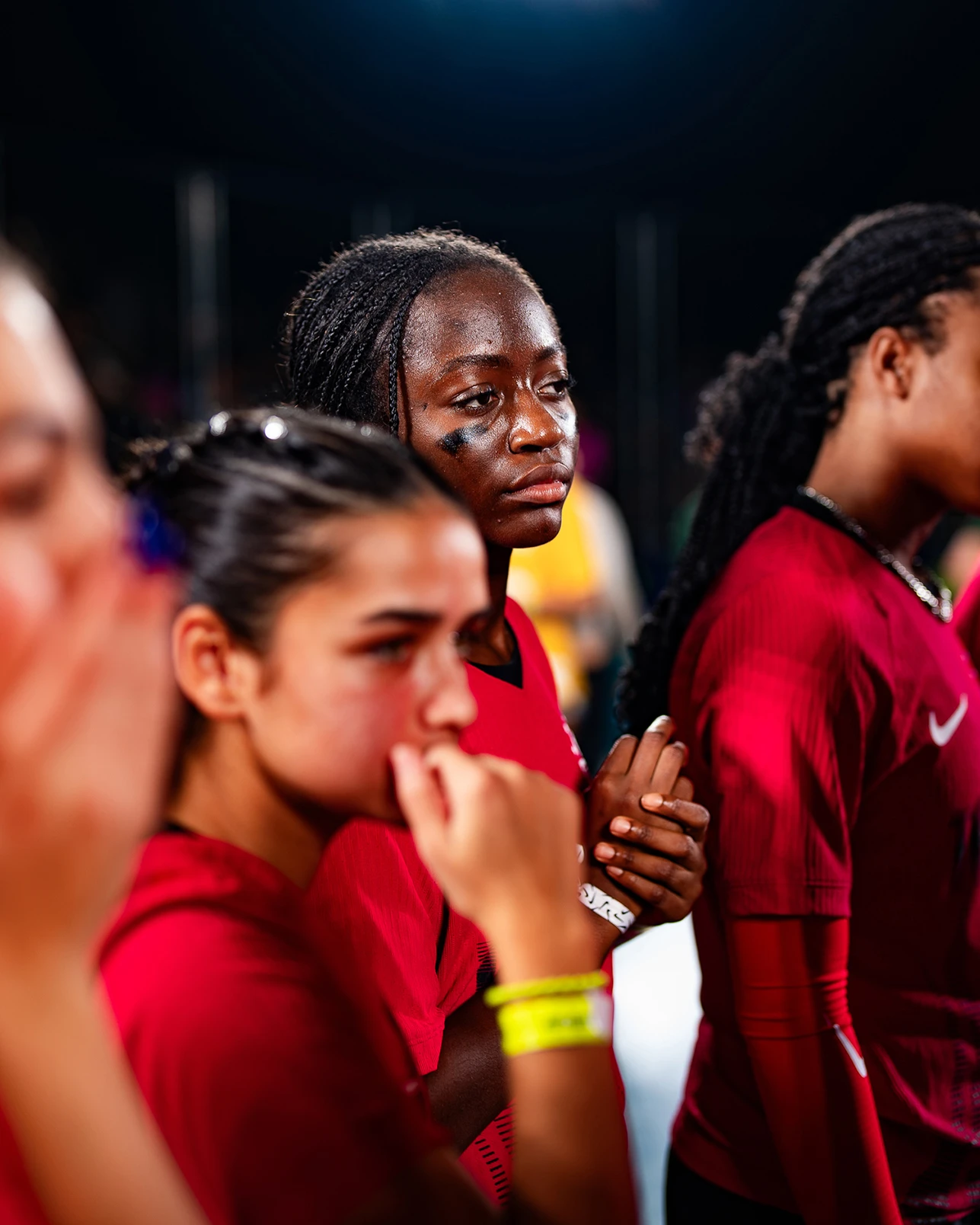 A group of athletes in red uniforms stands close together, looking serious and contemplative under bright indoor lighting.