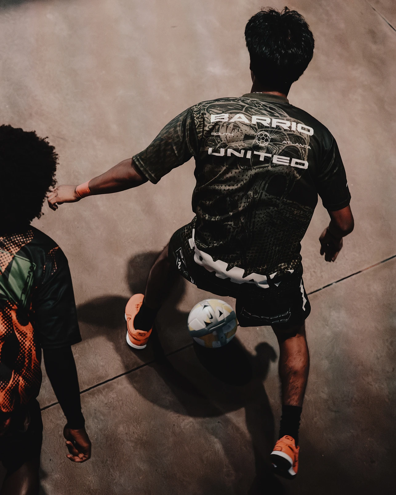 Two people play indoor soccer; one player in a “BARRIO UNITED” jersey controls the ball with their foot, while another player approaches from the left.