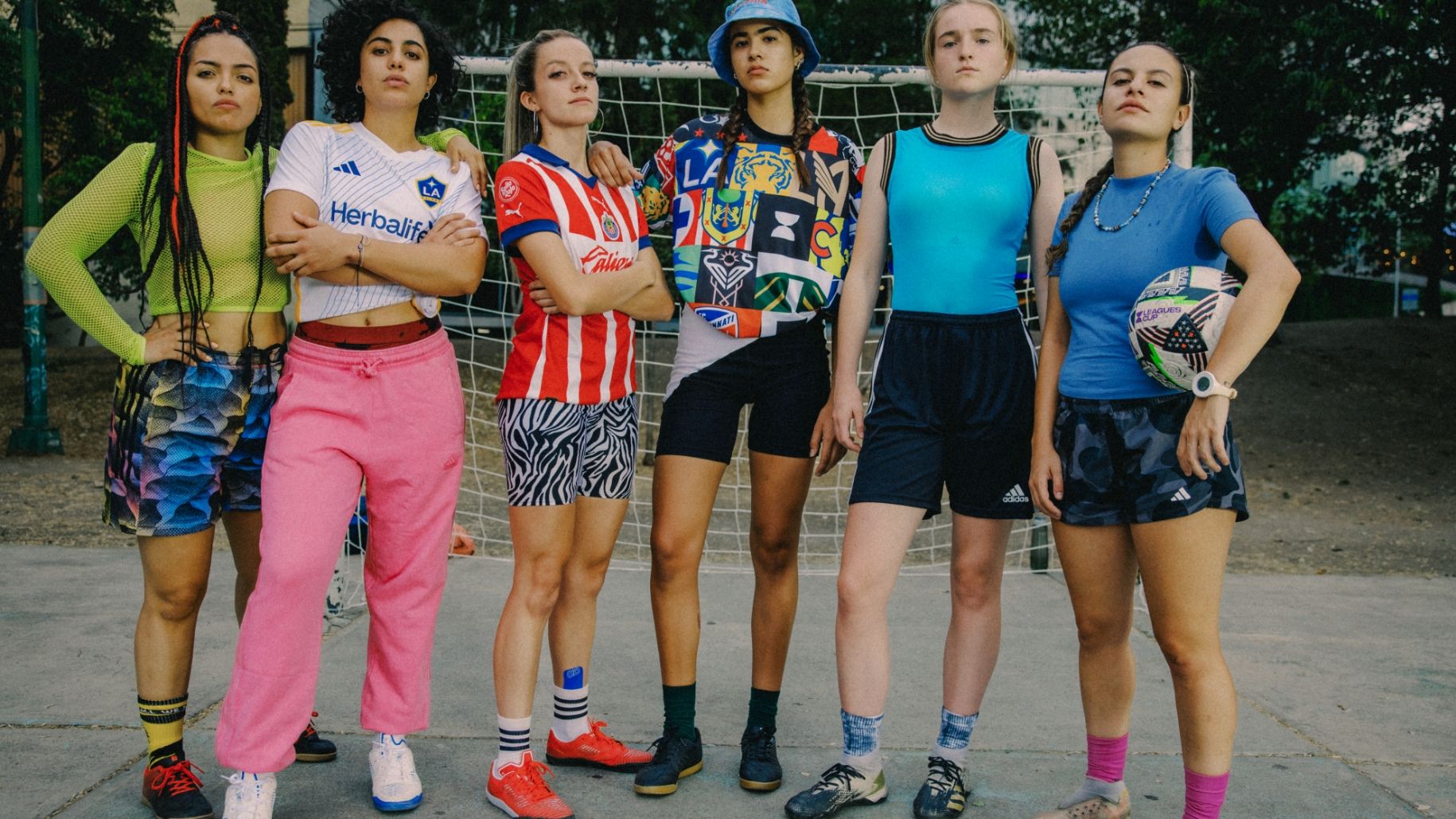 Six women in diverse sports attire stand confidently in front of a soccer goal, holding a soccer ball and exuding a sense of teamwork and determination.