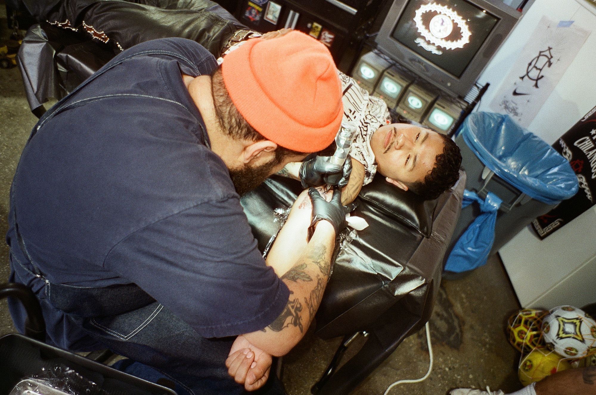 A tattoo artist in a beanie and gloves tattoos the forearm of a person lying on a black chair in a studio with various equipment and decor.