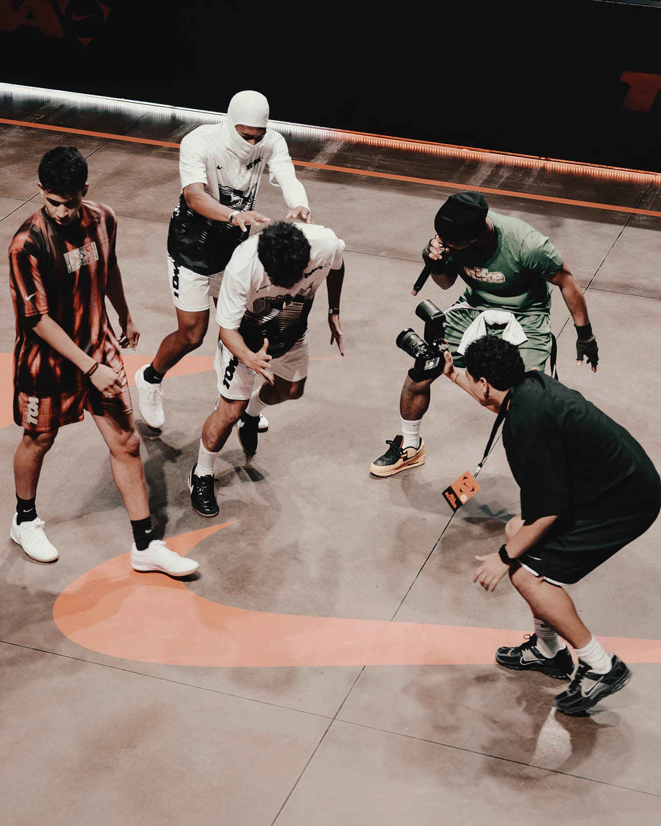 Five men interact on an indoor court, with two holding cameras and three appearing ready to play or pose. An orange swoosh is visible on the floor.