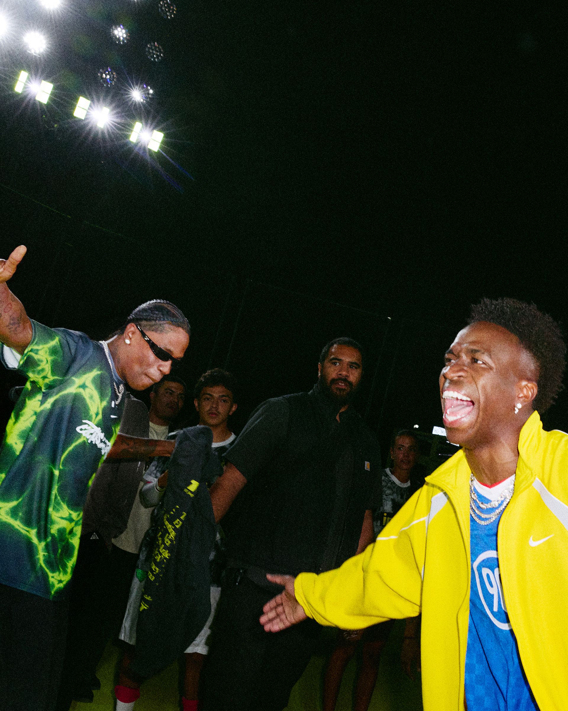 Two men gesture energetically towards each other on a lit stage, surrounded by a crowd and security guard, with a nighttime setting in the background.