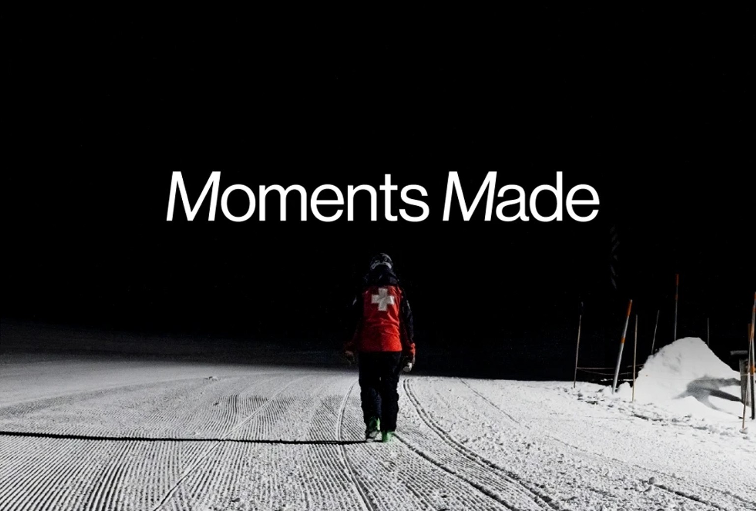 A person in a red jacket with a white cross walks alone on a snow-covered path at night; text above reads "Moments Made.